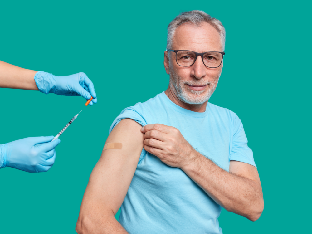 A middle-aged man wearing a T-shirt rolls up his sleeve to receive a vaccination. A healthcare professional wearing blue gloves prepares a syringe and holds a small vial. A plaster is visible on the man’s upper arm, suggesting he has just been vaccinated.
