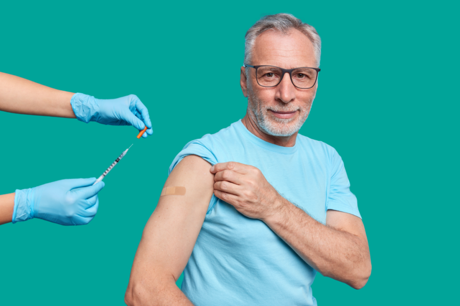 A middle-aged man wearing a T-shirt rolls up his sleeve to receive a vaccination. A healthcare professional wearing blue gloves prepares a syringe and holds a small vial. A plaster is visible on the man’s upper arm, suggesting he has just been vaccinated.