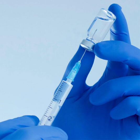 A pair of hands in blue surgical gloves holding needle and vial
