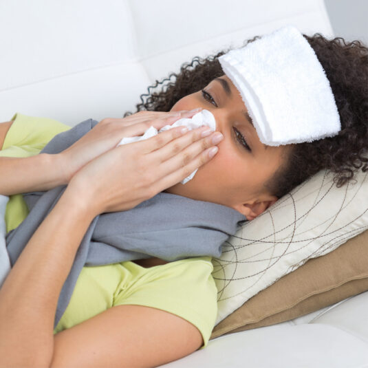 Unwell woman blowing nose and wearing a cold compress on forehead