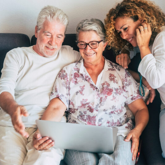 Older adults and a caregiver smiling while using a laptop together at home.