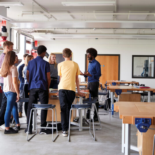 A teacher speaking to a diverse group of students standing in a classroom or workshop space.
