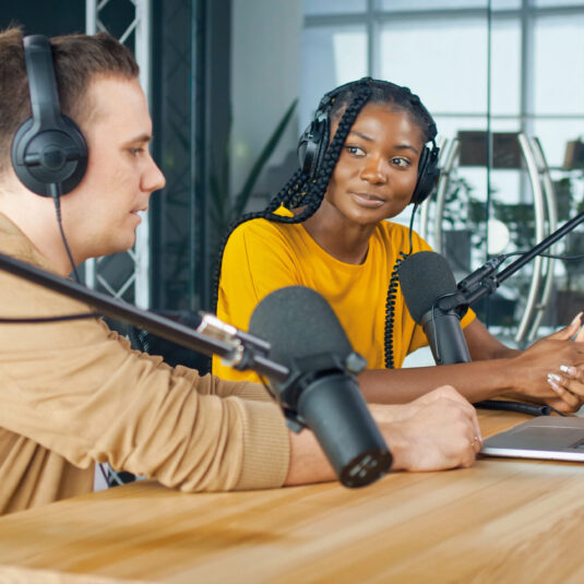 Two people wearing headphones speaking into microphones during a podcast or radio recording session.