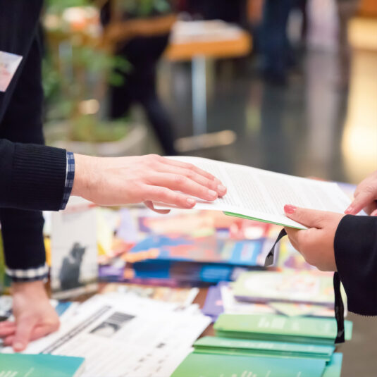 A person handing a leaflet or document to another person at an information table.