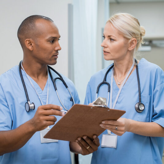 Two healthcare professionals in scrubs discussing information on a clipboard in a hospital or clinic setting.