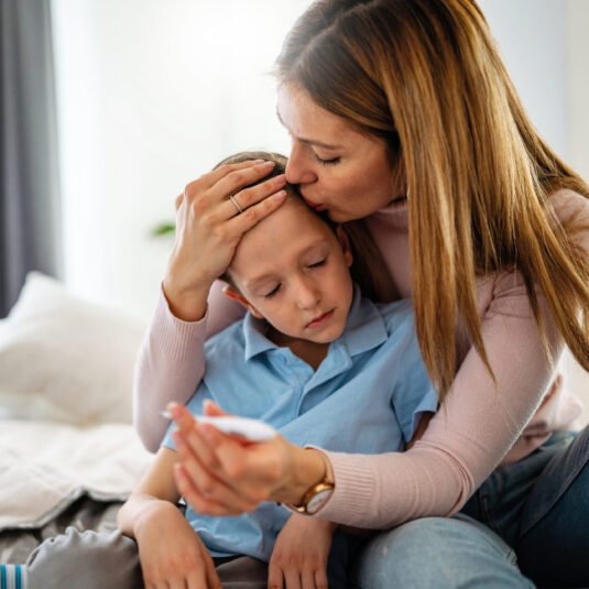 Mother checking her sick son’s temperature while comforting him on a bed.