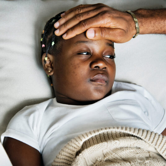 Parent checking a child’s forehead for fever while the child rests under a blanket.