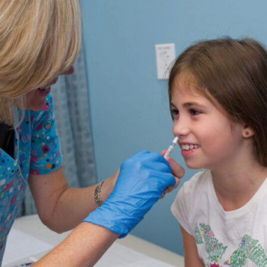 Healthcare worker administering a nasal vaccine spray to a smiling young girl.