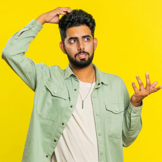 Confused man scratching his head while looking uncertain against a yellow background.