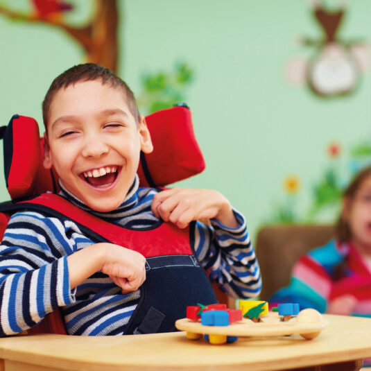Smiling child in a supportive seating chair playing with colourful toys.