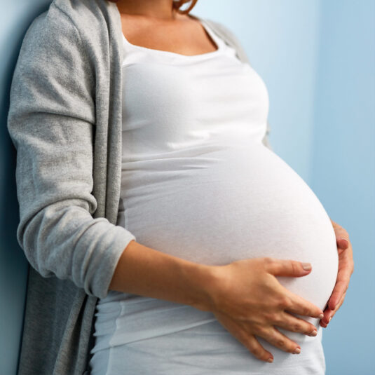 Pregnant woman holding her baby bump while leaning against a wall.