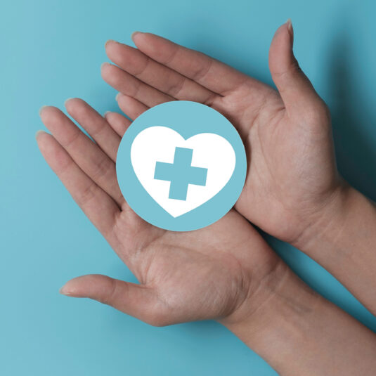 Hands holding a healthcare icon featuring a heart with a medical cross.
