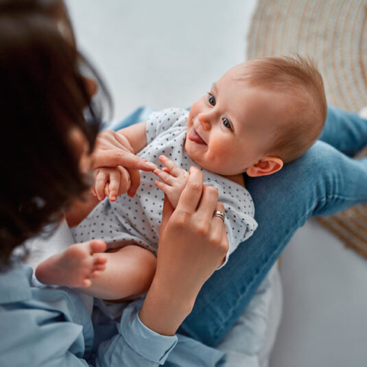 Smiling baby sitting on an adult’s lap while interacting playfully with their hands.