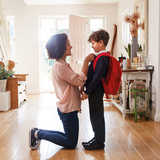 Parent kneeling to help a young child adjust their school uniform and backpack before school.