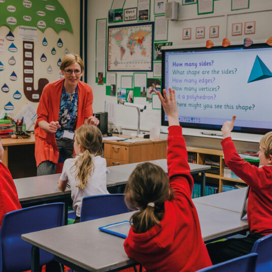 Teacher leading a classroom lesson while students raise their hands to answer questions.