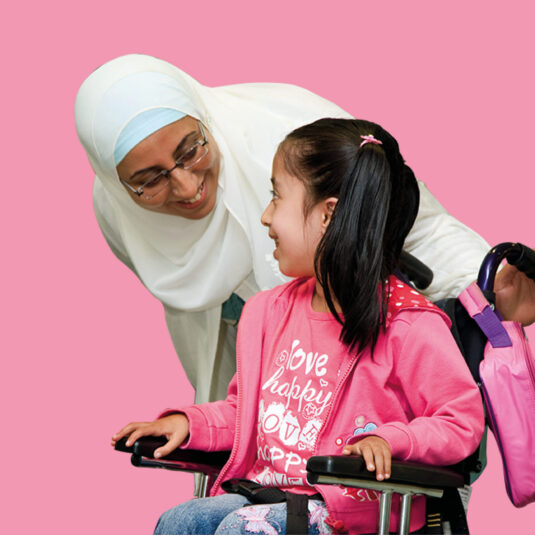 Woman smiling and interacting with a young girl sitting in a wheelchair, suggesting support, care, and inclusion.