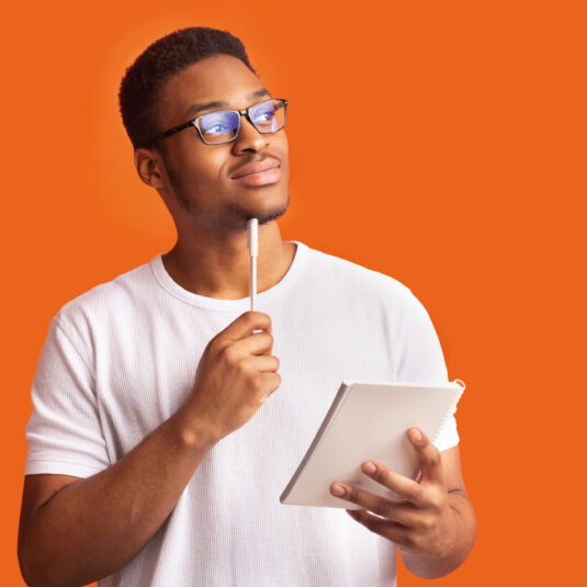 Young man wearing glasses, holding a notebook and pen while looking thoughtful, suggesting planning, reflection, or decision-making.