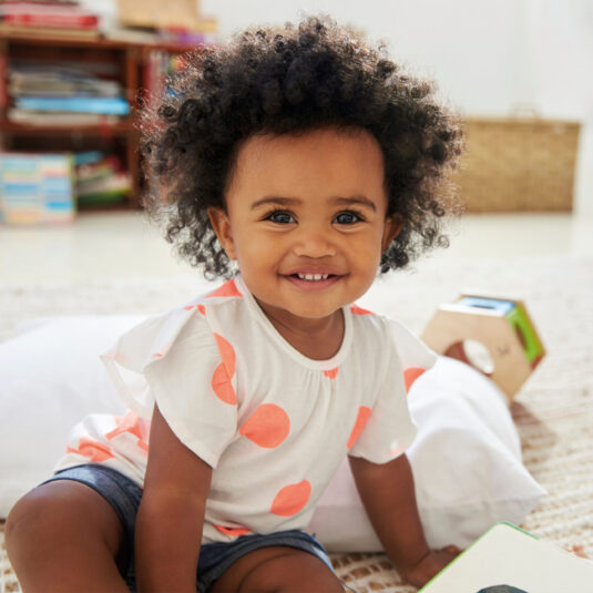 Smiling toddler with curly hair sitting indoors, appearing happy, healthy, and playful.