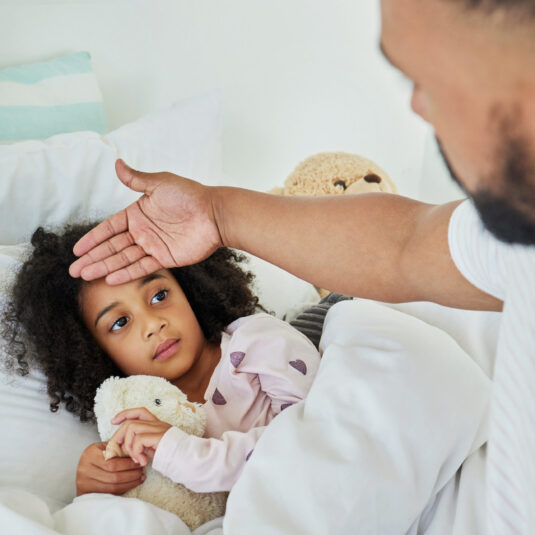 Parent checking a young girl’s temperature while she rests in bed holding a teddy bear, suggesting illness or fever monitoring.