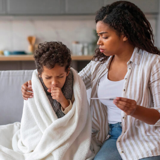 Mother comforting a sick child wrapped in a blanket while checking a thermometer, suggesting cold or flu symptoms.