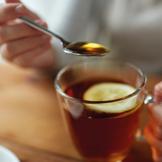 Close-up of a spoonful of honey above a cup of hot tea with lemon, representing a home remedy for sore throat or cough relief.