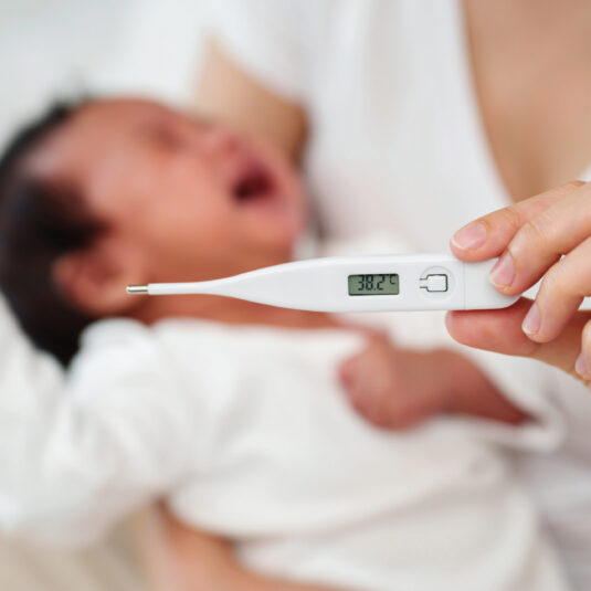 Digital thermometer showing a high temperature beside a newborn baby, indicating fever in an infant.