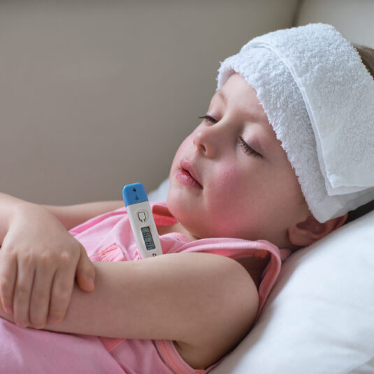 Young child lying in bed with a thermometer and cool cloth on the head, representing fever management and rest during illness.