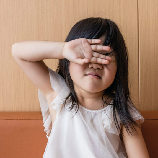 Young girl shielding her eyes and appearing unwell, suggesting tiredness, sensitivity, or feeling sick during illness.