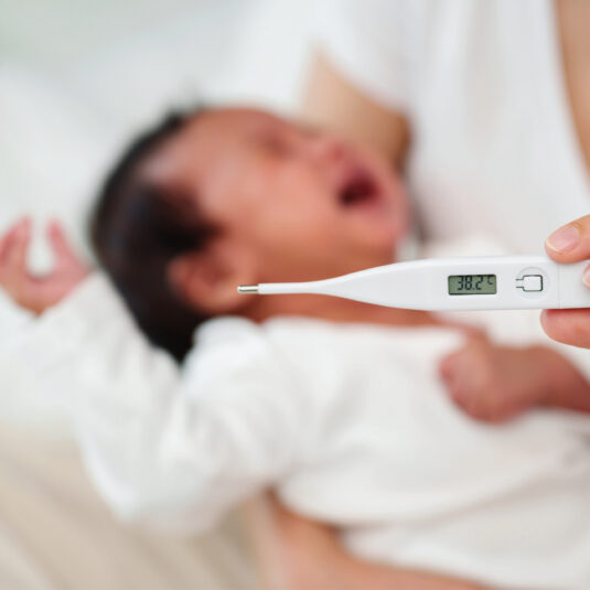 Digital thermometer showing a temperature of 38.2°C beside a blurred baby, indicating fever in an infant.