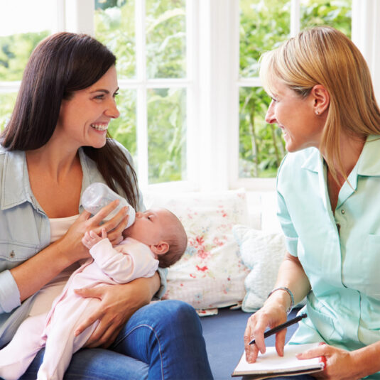 Health visitor or nurse speaking with a mother feeding her baby, representing family support and infant care guidance at home.