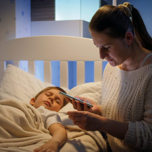 Parent checking on an unwell child at night while holding a phone and thermometer, suggesting monitoring symptoms or seeking advice.